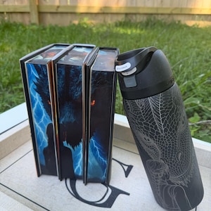 May include: A black insulated water bottle with a dragon design next to three books with fantasy art covers. The books have images of silhouettes, lightning, and a dragon. The water bottle has a black lid with a button that says "Lift to Unlock".