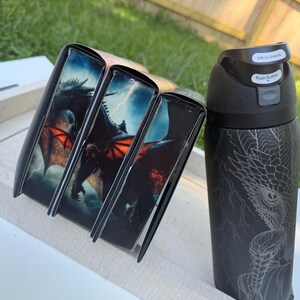 K&ouml;nnte beinhalten: Eine schwarze Wasserflasche und drei schwarze Buchh&uuml;llen. Die H&uuml;llen zeigen eine Drachenillustration mit roten Fl&uuml;geln vor einem st&uuml;rmischen Himmel. Die Wasserflasche hat ein Drachendesign und den Text "Lift to Unlock" und "Push Button".