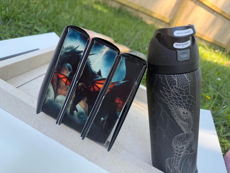 K&ouml;nnte beinhalten: Eine schwarze Wasserflasche und drei schwarze Buchh&uuml;llen. Die H&uuml;llen zeigen eine Drachenillustration mit roten Fl&uuml;geln vor einem st&uuml;rmischen Himmel. Die Wasserflasche hat ein Drachendesign und den Text "Lift to Unlock" und "Push Button".