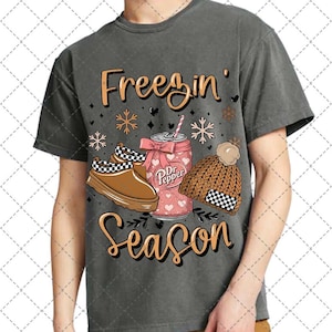 May include: Dark gray t-shirt with a graphic design featuring the words "Freezin' Season" in gold script. The design includes a Dr. Pepper can, a pair of brown clogs, and a knit hat with a checkered pattern.