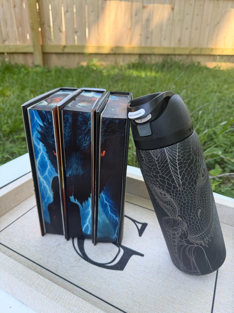 K&ouml;nnte beinhalten: Eine schwarze Wasserflasche mit einem Drachendesign und drei B&uuml;chern mit Fantasy-Kunst auf den Einb&auml;nden. Die B&uuml;cher stehen aufrecht neben der Wasserflasche. Die Wasserflasche hat einen schwarzen Deckel mit einem Knopf mit der Aufschrift "Lift to Unlock".