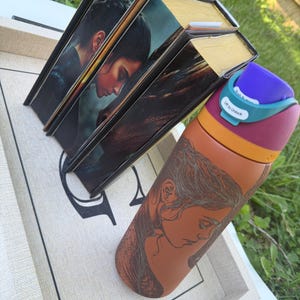 May include: Three hardcover books with gold-colored edges and a portrait of a person on the cover. A water bottle with a woman's portrait is next to the books. The water bottle has a purple lid and a teal-colored lock.