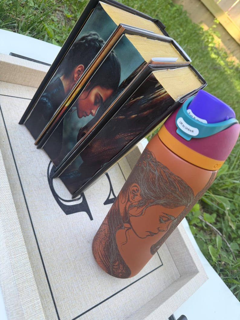 May include: Three hardcover books with gold-colored edges and a portrait of a person on the cover. A water bottle with a woman's portrait is next to the books. The water bottle has a purple lid and a teal-colored lock.