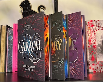 May include: A bookshelf displays a collection of hardcover books, including "Caraval" by Stephanie Garber. A black cat-shaped decoration sits atop the books. The books have colorful covers with intricate designs and titles. The shelf is black, and the background is blurred.