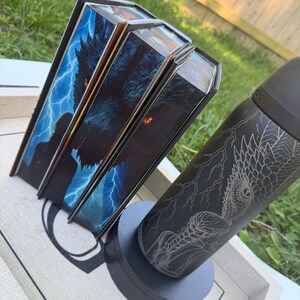 K&ouml;nnte beinhalten: Drei Buchboxen mit einem Drachen- und Blitzdesign und ein schwarzer Becher mit einem Drachen- und Blitzdesign. Die Buchboxen haben ein blaues und schwarzes Farbschema. Der Becher steht auf einem grauen Sockel.