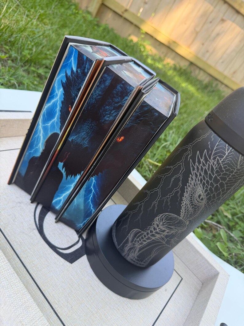 K&ouml;nnte beinhalten: Drei Buchboxen mit einem Drachen- und Blitzdesign und ein schwarzer Becher mit einem Drachen- und Blitzdesign. Die Buchboxen haben ein blaues und schwarzes Farbschema. Der Becher steht auf einem grauen Sockel.