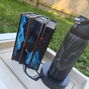 May include: Three book boxes with a fantasy theme, featuring a wolf and lightning design, are displayed on a tray. A black water bottle with a dragon engraving sits on a black base, next to the books. The background is a grassy area.