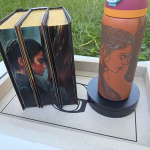 May include: A set of three books with gold-colored edges and a water bottle with a woman's portrait on a tray. The books have a portrait of a woman on the cover. The water bottle is orange and has a pink lid.