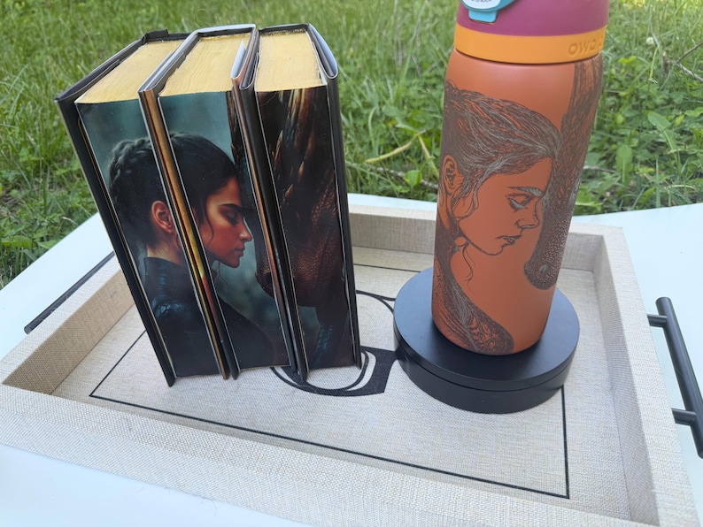 May include: A set of three books with gold-colored edges and a water bottle with a woman's portrait on a tray. The books have a portrait of a woman on the cover. The water bottle is orange and has a pink lid.