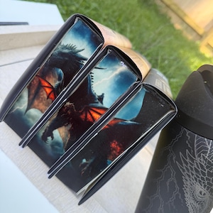 May include: Several books with black spines and dragon illustrations. The dragon has red wings and is set against a stormy sky. A black tumbler with a dragon design is also visible.