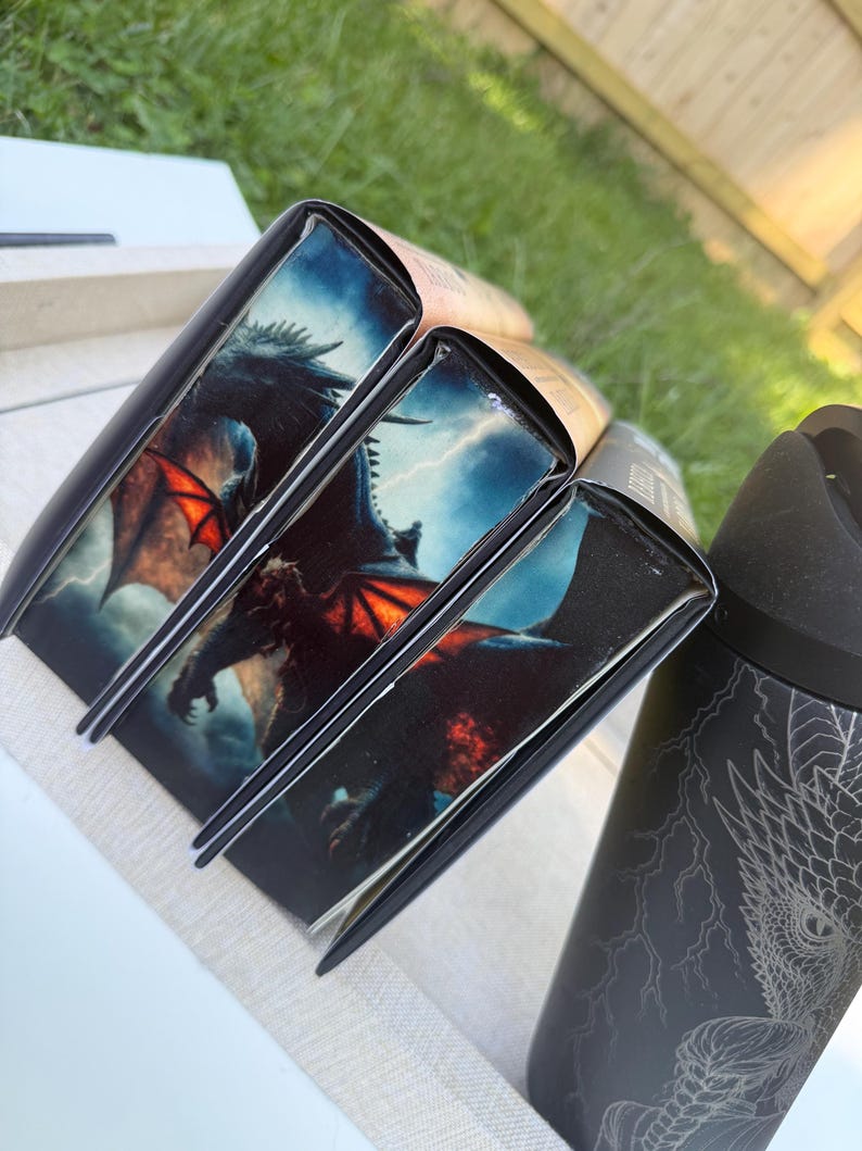 May include: Several books with black spines and dragon illustrations. The dragon has red wings and is set against a stormy sky. A black tumbler with a dragon design is also visible.