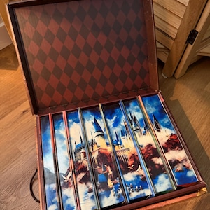 Puede incluir: Maleta marrón abierta con un patrón de diamantes rojos y negros en el interior. En su interior hay varias cajas rectangulares con imágenes de un castillo en una montaña, sobre un cielo azul con nubes.
