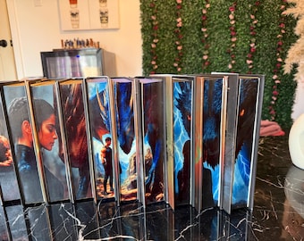 May include: A collection of hardcover books with fantasy art covers. The spines are visible, showcasing vibrant illustrations of dragons, figures, and landscapes. The books are arranged on a glossy black surface.