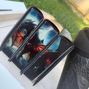 K&ouml;nnte beinhalten: Ein Stapel B&uuml;cher mit schwarzen Buchr&uuml;cken und Drachenillustrationen auf den Einb&auml;nden. Die Drachenillustration zeigt einen dunklen Drachen mit roten Fl&uuml;geln vor einem st&uuml;rmischen Himmel. Ein schwarzer Becher mit einem Drachendesign ist ebenfalls zu sehen.