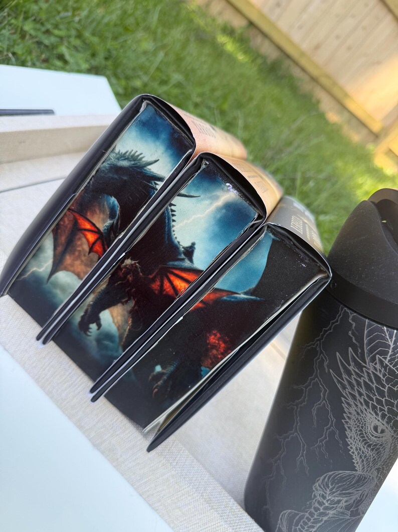 K&ouml;nnte beinhalten: Ein Stapel B&uuml;cher mit schwarzen Buchr&uuml;cken und Drachenillustrationen auf den Einb&auml;nden. Die Drachenillustration zeigt einen dunklen Drachen mit roten Fl&uuml;geln vor einem st&uuml;rmischen Himmel. Ein schwarzer Becher mit einem Drachendesign ist ebenfalls zu sehen.