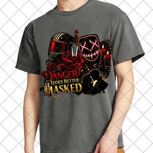 Puede incluir: Camiseta gris con un diseño gráfico que incluye un casco de moto rojo y negro, una daga, una máscara brillante, rosas y velas. El texto "DANGER LOOKS BETTER MASKED" está en rojo y dorado.