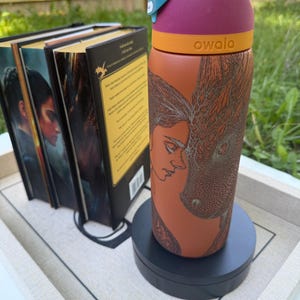 May include: A colorful Owala water bottle with a dragon and woman design. The bottle has a purple top, orange band, and a rust-colored body. The bottle is on a black stand, with a few books in the background.