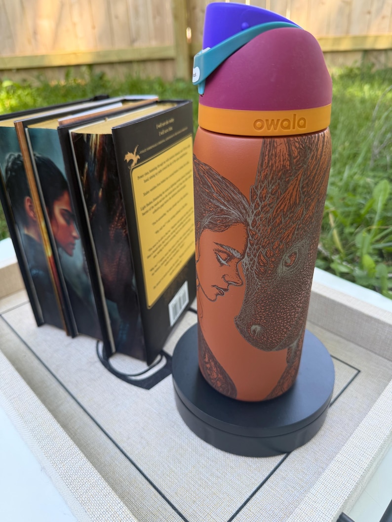 May include: A colorful Owala water bottle with a dragon and woman design. The bottle has a purple top, orange band, and a rust-colored body. The bottle is on a black stand, with a few books in the background.