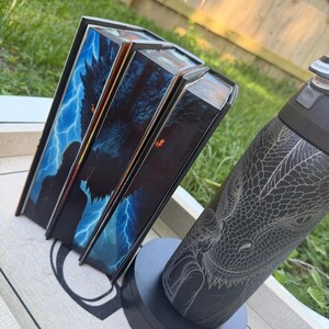 K&ouml;nnte beinhalten: Drei Hardcover-B&uuml;cher mit Fantasy-Motiven, die einen Drachen und Blitze auf den Einb&auml;nden zeigen. Eine schwarze Wasserflasche mit Drachengravur ist ebenfalls vorhanden und steht auf einem grauen St&auml;nder.