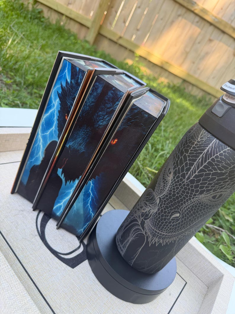 K&ouml;nnte beinhalten: Drei Hardcover-B&uuml;cher mit Fantasy-Motiven, die einen Drachen und Blitze auf den Einb&auml;nden zeigen. Eine schwarze Wasserflasche mit Drachengravur ist ebenfalls vorhanden und steht auf einem grauen St&auml;nder.