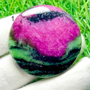 Op de afbeelding: Een ronde, gepolijste cabochon edelsteen met een levendig magenta-roze centrum, omringd door banden van diepgroene en zwarte minerale insluitingen. De steen heeft een glad, reflecterend oppervlak en is gemarkeerd met het cijfer 1.