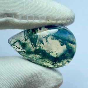 May include: A teardrop-shaped moss agate gemstone, held between two white fabric-covered fingers. The translucent stone features green, blue, and white inclusions, creating a natural, organic pattern. The stone is polished and smooth.