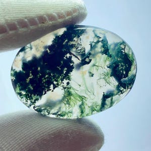 May include: An oval-shaped moss agate gemstone. The translucent stone features green, white, and dark green inclusions that resemble a miniature landscape. The stone is held between two fingers covered in a white glove.