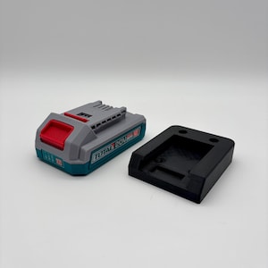 Peut inclure: Un bloc-batterie TOTAL 20V gris et turquoise avec un dessus rouge, à côté d'un support de batterie noir. Le bloc-batterie porte le nom de la marque.