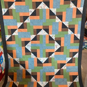 May include: A handmade quilt with a geometric pattern. The quilt features squares and triangles in shades of blue, green, orange, black, and brown. The quilt has a dark gray border and is likely made of cotton fabric.