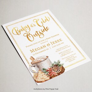 May include: A white rectangular baby shower invitation with a gold foil border and gold text reading Baby it's Cold Outside. It features a watercolor illustration of a mug with marshmallows, a pinecone, and a decorated cookie.