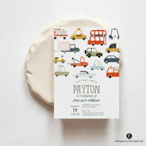 May include: A white invitation card featuring various cartoon vehicles in orange, yellow, blue, and green. The text "PAYTON IS TURNING 21" is displayed, along with event details. The card is on a white, textured surface.