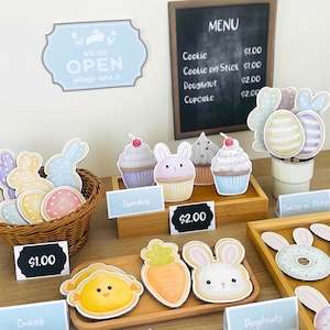 May include: A display of Easter-themed treats, including cookies, cupcakes, and doughnuts, arranged on wooden trays. The treats are shaped like bunnies, eggs, and carrots. A chalkboard menu lists prices. A sign says "We're Open".