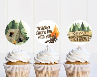 May include: Three round cupcake toppers with a camping theme. The toppers feature a green and brown tent, a campfire with orange flames, and a wooden sign that says "Camp Matthews".