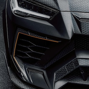 May include: Close-up of a black luxury car's front end. The car features a textured hood, a sleek headlight design, and a black grille with orange accent lines. The overall aesthetic is modern and aggressive.