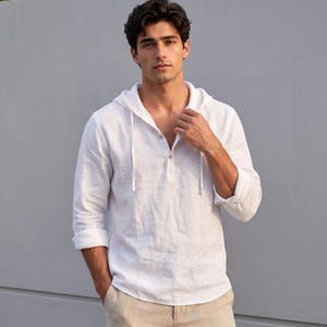 May include: A white linen hooded shirt with long sleeves and a button-down neckline. The shirt features a drawstring hood and is worn with beige shorts. The model is standing in front of a grey wall.