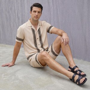 May include: A beige two-piece matching set featuring a short-sleeved button-down shirt and shorts. The shirt and shorts have a dark brown decorative pattern. The person is wearing black sandals.