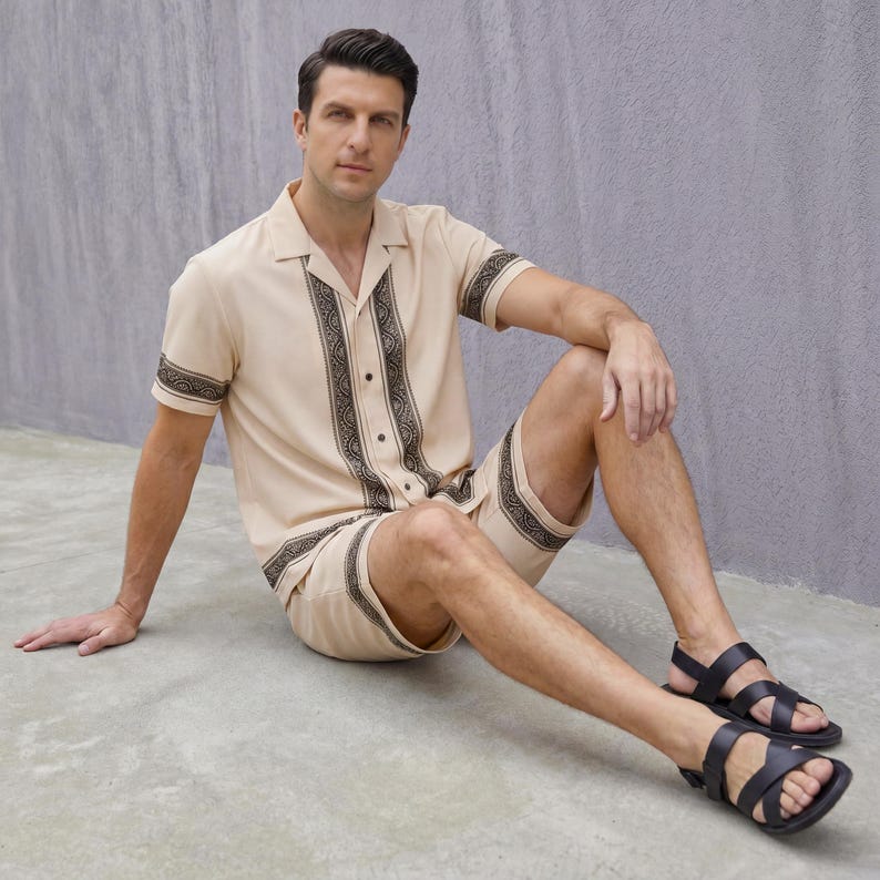 May include: A beige two-piece matching set featuring a short-sleeved button-down shirt and shorts. The shirt and shorts have a dark brown decorative pattern. The person is wearing black sandals.