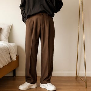 May include: Brown pleated trousers with a high waist. The pants are paired with a black top and white sneakers. A gold floor lamp stands in the background, next to a bed with a brown pillow and white bedding.