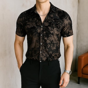 May include: Black and brown short-sleeve button-down shirt with a floral pattern. The shirt has a classic collar and is paired with black pants. A silver watch and chain are visible.