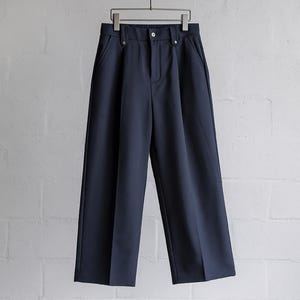 May include: Navy blue wide-leg pants hanging on a metal hanger against a white brick wall. The pants have a button and zipper closure, with two front pockets. The fabric appears to be a smooth, woven material.