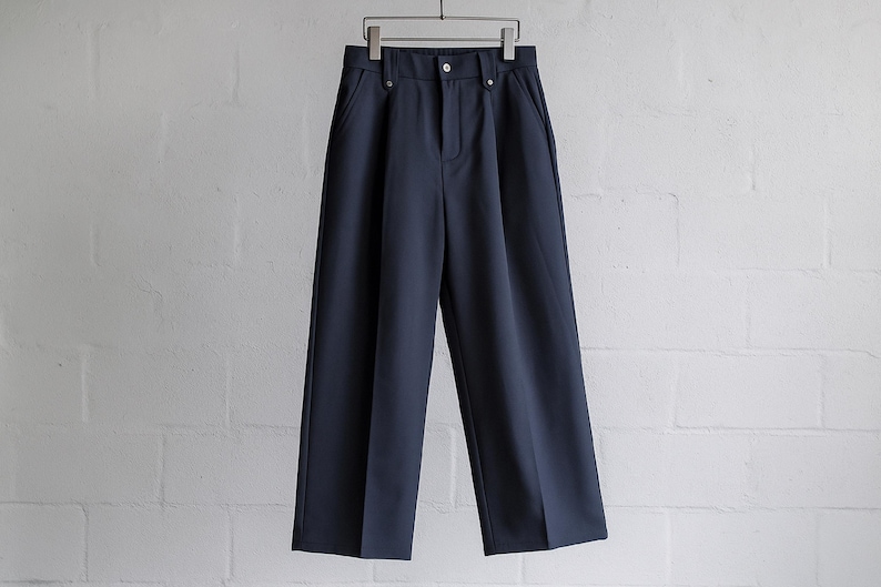 May include: Navy blue wide-leg pants hanging on a metal hanger against a white brick wall. The pants have a button and zipper closure, with two front pockets. The fabric appears to be a smooth, woven material.