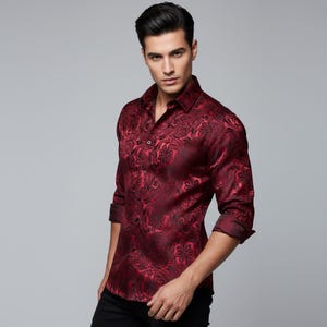 May include: A burgundy button-down shirt with a dark paisley pattern. The long-sleeved shirt has a classic collar and is made of a silky material. The sleeves are rolled up, revealing the dark burgundy cuffs. The shirt is worn with black pants.