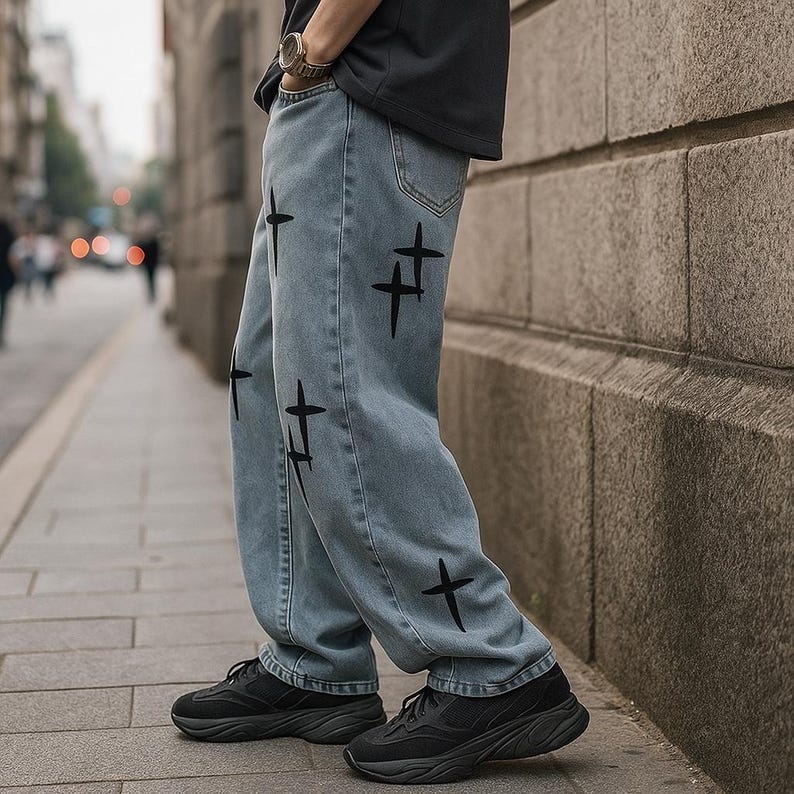 May include: Light blue denim jeans featuring black cross patterns. The jeans have a loose fit and are worn with black trainers. The image shows a person standing on a pavement.
