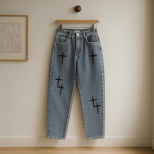 May include: Pale blue denim jeans featuring black cross and '4' designs. The jeans are displayed on a wooden hanger. They have a classic five-pocket design and a button and zip closure. The background is a neutral wall.