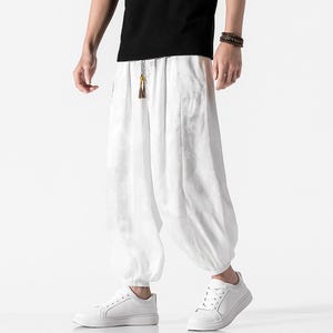 May include: White, loose-fitting pants with a subtle floral pattern. The pants have a drawstring waist and are gathered at the ankles. The person is wearing white sneakers and a black shirt. The pants appear to be made of a lightweight fabric.