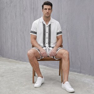 May include: A white short-sleeved shirt and matching shorts set with black decorative patterns. The shirt has a collared neckline and button-up front. The shorts have a drawstring waist. The person is wearing white sneakers.