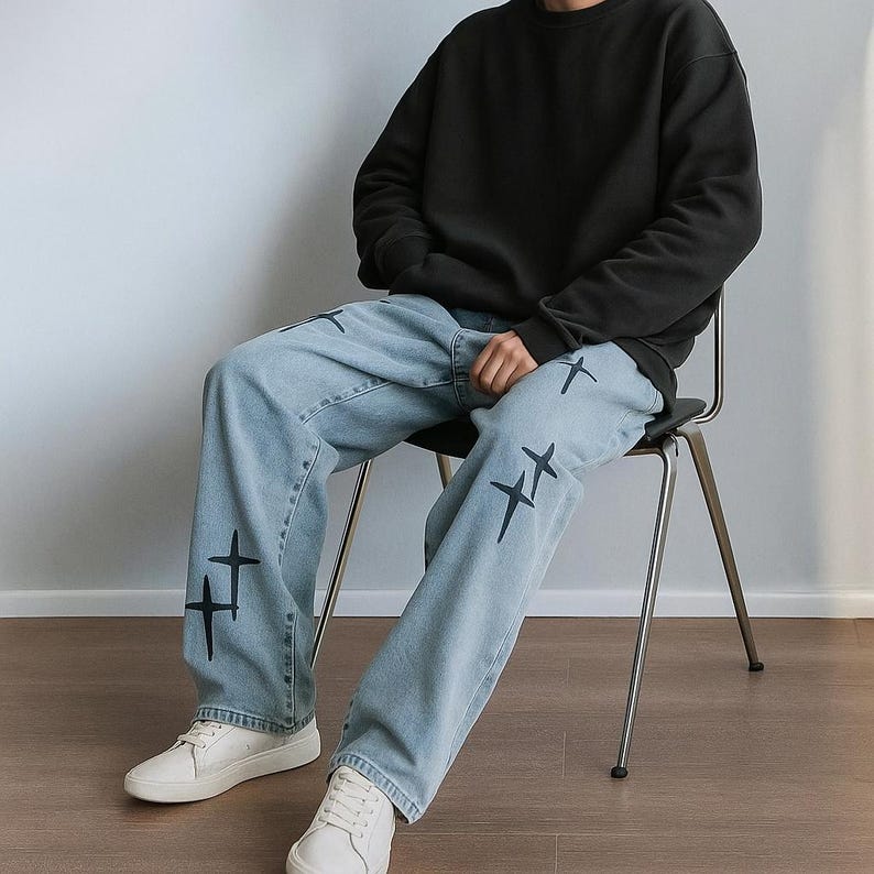 May include: A person seated wearing a black sweatshirt and light blue jeans. The jeans feature black cross designs. The outfit is completed with white trainers. Casual wear.