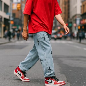 May include: Light blue denim jeans with black cross graphics. The person is wearing a red t-shirt and red, white, and black trainers with black laces. The trainers have a white Nike swoosh.