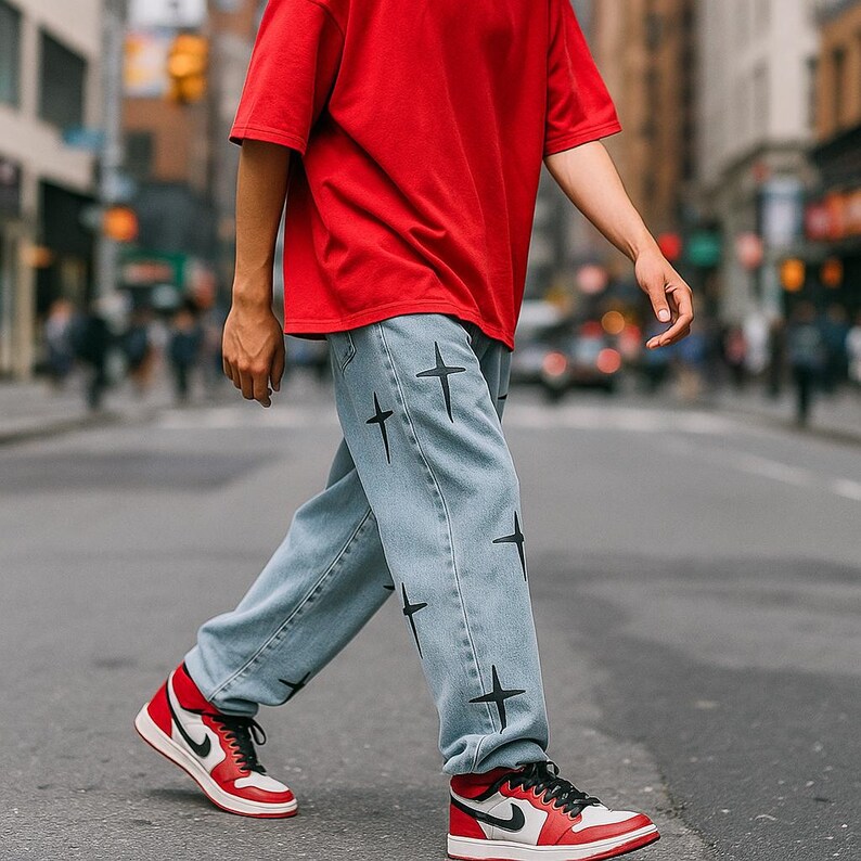 May include: Light blue denim jeans with black cross graphics. The person is wearing a red t-shirt and red, white, and black trainers with black laces. The trainers have a white Nike swoosh.