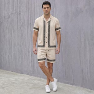 May include: A beige two-piece outfit consisting of a short-sleeved button-up shirt and matching shorts. The shirt and shorts have a black decorative border. The person is wearing white sneakers.